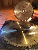 Photo showing two circular saw blades placed on a wooden surface. Larger 10 inch blade underneath and smaller 7 inch blade held above showing size difference and condition.