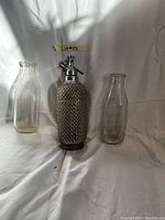 Full view of the antique Sparklets mesh glass siphon bottle with two clear milk bottles on either side, shown on white background.