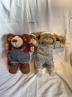 Front view of Cabbage Patch Kids doll and Furskins Bear plush sitting upright showing clothing and details