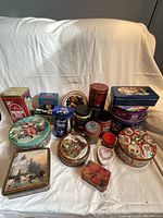 Full view of assorted collectible tins arranged on a white backdrop. Various shapes and colors including red, blue, green, and decorative motifs seen clearly.