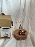 Photo of the handcrafted wooden music box on white background next to the lot number sticker. Shows wood base with Santa, sleigh, reindeer, and tree figurines.