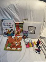 Photo showing various children's books, a plush rabbit pillow, a white decorative canvas with heart, colorful rubber bands, scissors, and craft supplies.