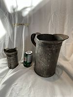 Front view of two metal jugs with soda can for scale. Larger jug approximately gallon size, smaller jug approximately pint size.