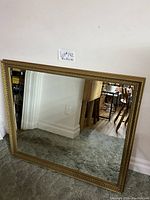 Rectangular framed mirror with gold decorative frame leaning against wall on carpeted floor. Frame has beaded and ornate detail along edges. Reflection shows part of a room with wood floor and furniture.