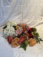 Photo showing a mixed collection of artificial flowers including hydrangeas, roses, daisies, and greenery arranged together on a white fabric background.