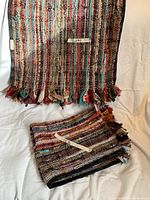 Photo shows two folded multi-color woven area rugs with vertical stripes and tassel edges lying on a white surface, clearly showing fabric texture and colors.