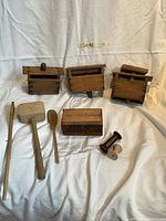 Photo of very old wooden kitchen tools including three butter molds, a butter holder box, spoon, butter paddle, long stick, and two spools on a white cloth background.