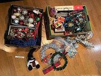 Top-down photo showing two boxes filled with vintage Christmas decorations including glass ornaments, ribbons, tinsel, wreaths, lights, and figurines