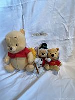 Photo shows three Winnie the Pooh bear plush toys of varying sizes, arranged against a white cloth backdrop.