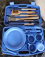 Blue carrying case opened, showing wooden and metal BBQ grilling tools on one side, blue plastic dinnerware on the other side.