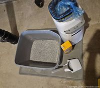 Photo showing white Litter Locker with blue refill bag, gray litter pan with litter, litter scoop, deodorizer box, and textured mat on floor.