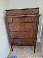 Front view of wood chest of drawers with 5 drawers, showing ornate hardware and turned legs with casters