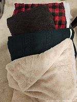Photo showing four blankets stacked on a floor: a red and black buffalo plaid blanket, dark brown plush blanket, dark green knitted blanket with white trim, and beige faux fur blanket.