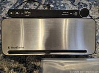 Top view of FoodSaver vacuum sealer showing stainless steel surface and black control panel with buttons and cord.