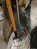 Photo showing several metal garden shovels with wooden handles along with two metal leaf rakes with different handles positioned on a floor.