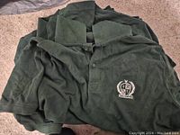 Stack of dark green golf shirts spread out showing button-down collar and crest embroidery on the chest.