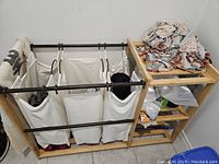 Laundry sorter showing white canvas bags hung on dark metal rods with wooden frame and shelving unit beside it containing folded fabrics and items.