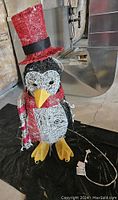 Front view of penguin ornament showing face, yellow beak, red top hat, red scarf, and part of the light string.