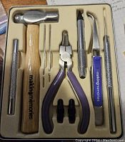 Scrapbooking tools including mini hammer with wood handle, pliers with purple grips, tweezers, blades, and needle tips arranged in fitted tray