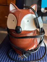 Photo of red ceramic fox-shaped wax melter with electrical cord wrapped around it, showing detailed facial colors and part of cord plug.