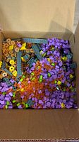 Box filled with purple, yellow, gray, and brown LEGO bricks and plates including minifigure parts and accessories.
