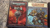 The photo shows two books, Warhammer 40,000 and Dungeons & Dragons Player's Handbook placed side by side on a carpeted surface.