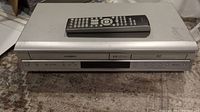 Front view of Toshiba DVD Recorder/VCR combo unit with remote on top, showing control buttons and disc slot.