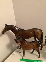 Two ceramic horse figurines including a large horse and a smaller pony, both brown and glossy, placed against a wall.