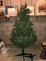 Front view of artificial Christmas tree with green PVC branches and black metal tripod stand base, captured indoors against a concrete block wall and tiled floor.