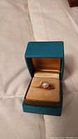 Ring displayed inside teal and beige box, showing overall design and pearl centerpiece.