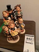 Front view of five vintage Hummel figurines grouped together on a table showing details and size relative to auction tag.