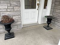 Pair of black cast iron planters positioned outside a house entrance on concrete flooring.