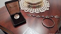 Overview of all jewelry items on wooden table with lace doily, showing the bracelet, bangle, and brooch in box.