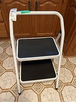 Front view of white folding metal step stool with two black steps with non-slip texture, placed on tiled floor in front of wooden cabinets.