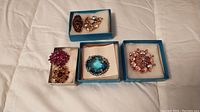 Overall view of six vintage brooches in individual boxes with cushions. Assortment of colorful rhinestones including pink, purple, amber, and blue.