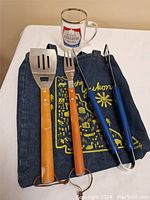 Three BBQ tools with wooden and blue handles, a branded beer mug, and a Yukon-themed cloth arranged on a white surface.