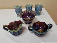 Photo showing creamer, sugar bowl, bowl, and three goblet glasses arranged on white surface, highlighting the iridescent blue carnival glass with embossed patterns.