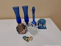 Photo showing all items in the lot arranged on white surface: two blue vases, tall blue bottle with stopper, blue bear plaque, clear glass bowl, round reflective plaque with floral design, and fish couple ceramic figurine.