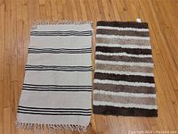 Two floor mats side by side on hardwood floor: beige woven mat with black stripes and fringed ends; shaggy striped mat in brown, cream, and tan colors