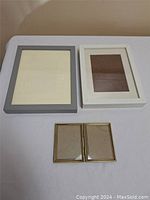 Four picture frames displayed on a white surface: one large grey frame, one large white frame, and two small gold-tone frames with decorative edges.