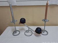 Three hand forged steel candle holders with spiral bases, two holding candles and center one featuring black spheres on copper bases connected by an arm.