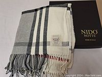 Folded Nido Notte Italian throw blanket beside its original box showing the throw's plaid pattern and fringed edges.