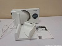 White Salton electric food slicer with attached cord and blue thickness dial, white plastic food holder, and instruction manual on white surface