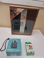 Photo showing the shadow box with wooden frame and glass front, packaged Instax Mini 11 camera box, and two packs of Instax Mini instant film.