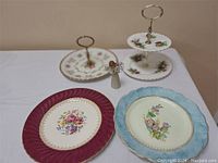 Four decorative plates and a two-tier plate stand displayed on a white cloth with salt and pepper shakers in the center, showing variety of floral patterns and colors