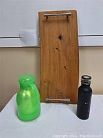 Wooden serving platter standing upright flanked by a green plastic bottle on the left and a black metal bottle on the right, showing full height and handles.