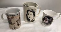 Three large coffee mugs featuring British Royalty images and commemorative texts photographed together on a dark cloth background.