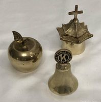Photo showing three brass hand bells: apple-shaped, pagoda-shaped with cross, and Kiwanis International insignia bell, positioned on a white background.