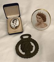 Set of three commemorative items: a brooch in a blue velvet-lined box, an oval china plaque, and a brass medallion with handle-like top.