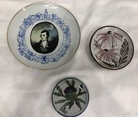Three plates laid flat including Robert Burns collector plate with blue design and gold rim, floral clay pottery plate, and small thistle stoneware coaster.